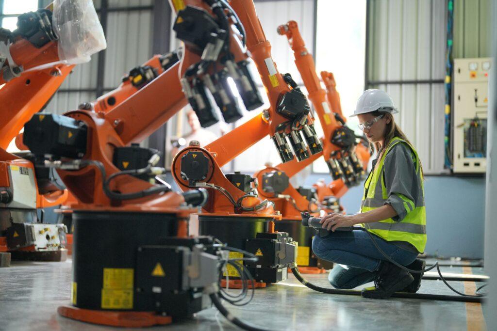 Eine Ingenieurin im Schutzhelm und Warnweste arbeitet an der Programmierung von orangefarbenen Industrierobotern in einer modernen Fabrikumgebung.