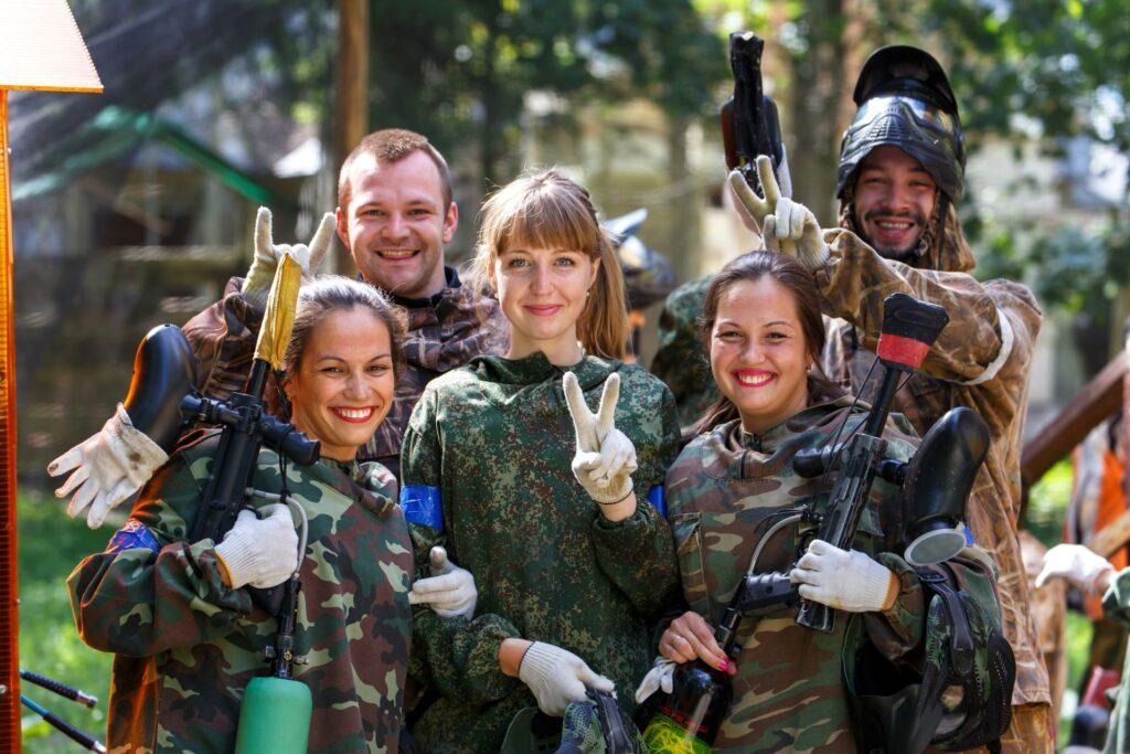 Gruppe von Paintball-Spielern in Tarnkleidung lächelt in die Kamera – Eine fröhliche Gruppe von fünf Paintball-Spielern in Tarnkleidung posiert nach einem Spiel und zeigt das Victory-Zeichen.