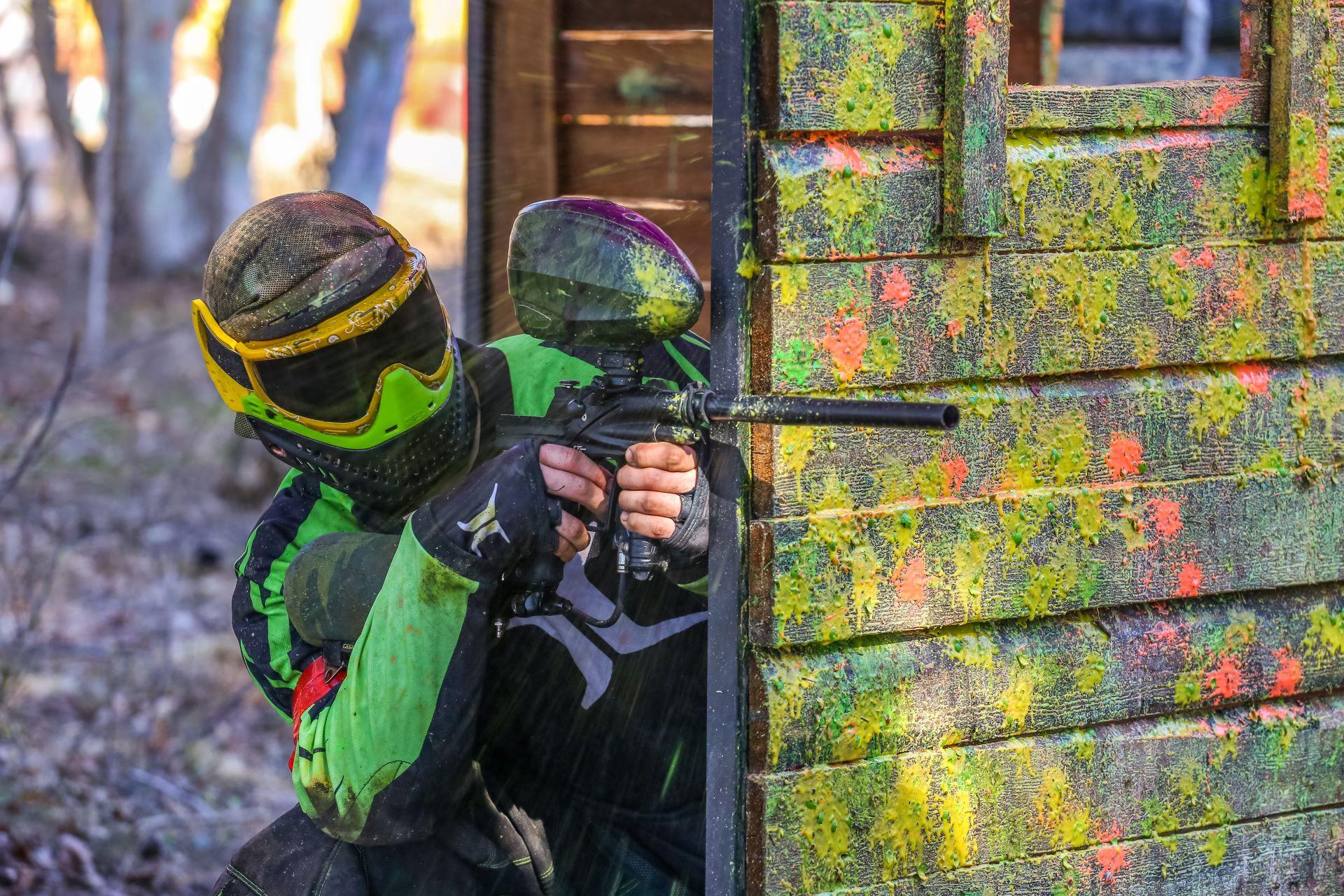 Paintball-Spieler hinter bunter Deckung – Ein Paintball-Spieler in grün-schwarzer Ausrüstung versteckt sich hinter einer von Farbkugeln getroffenen Wand und zielt auf den Gegner.