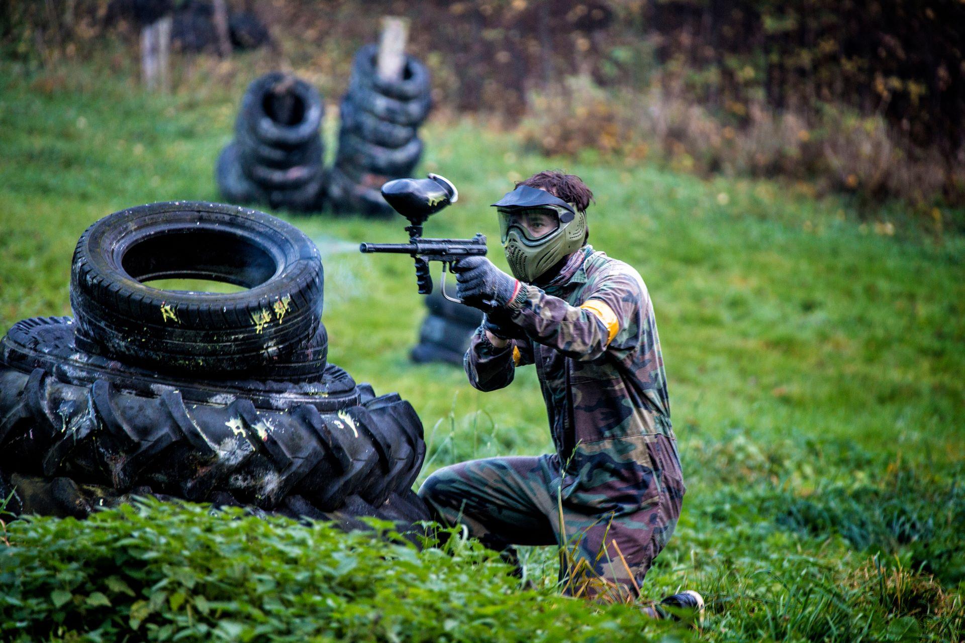 Person in Paintball-Ausrüstung hinter einem Reifenstapel in Deckung – Eine Person in Tarnkleidung und Schutzmaske zielt mit einem Paintball-Markierer hinter einem Stapel alter Reifen, während sie sich auf dem Spielfeld bewegt.