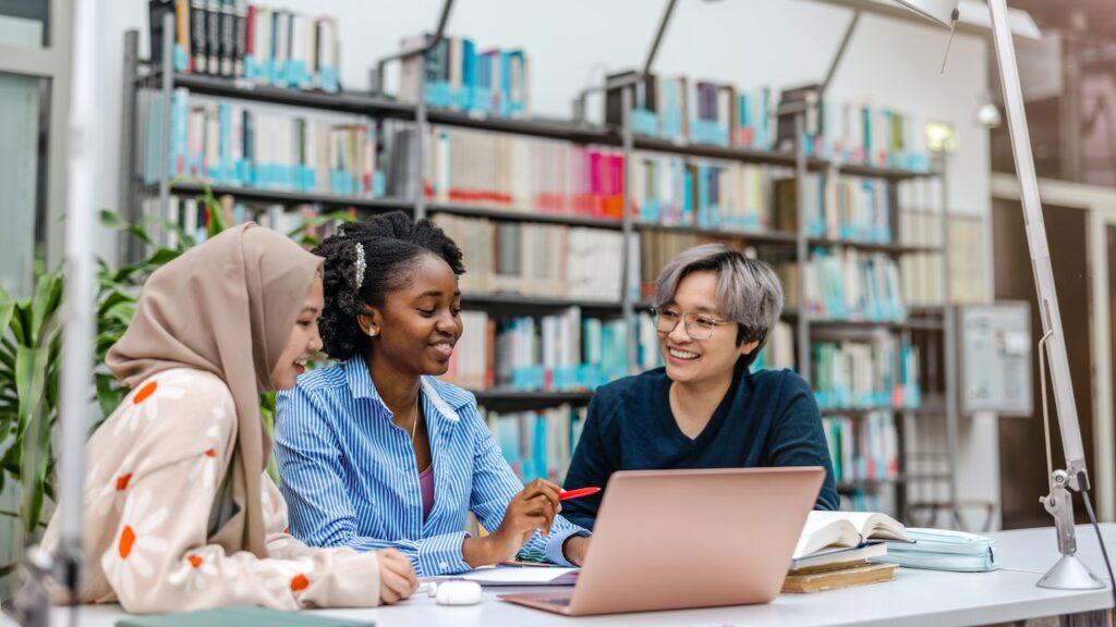 Drei Studierende verschiedener Kulturen arbeiten gemeinsam an einem Projekt in einer modernen Bibliothek.