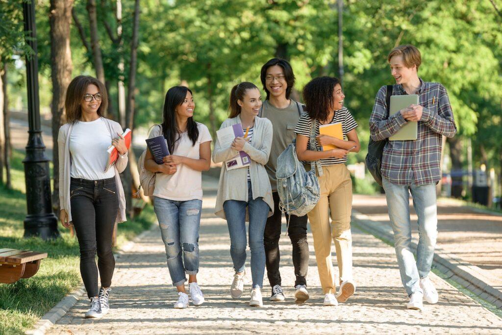 Gruppe internationaler Schüler spaziert lachend durch einen grünen Park mit Büchern und Rucksäcken in der Hand.