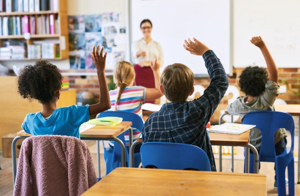 Schüler in einem multikulturellen Klassenzimmer heben die Hände, während eine Lehrerin vorne lächelt und Fragen beantwortet.